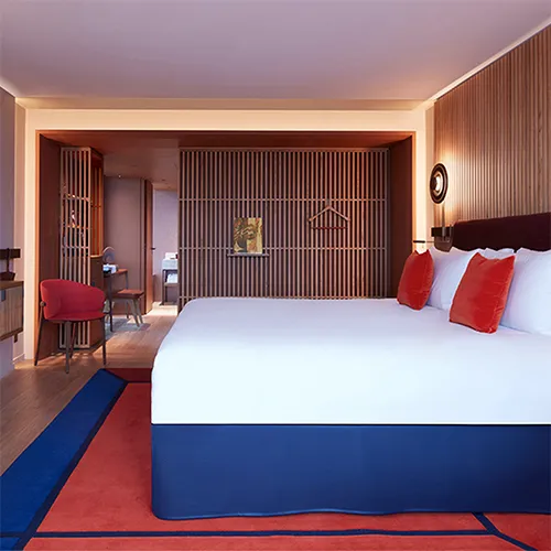 A modern hotel room with a large bed, white linens, red accent pillows, wooden panel walls, a red chair at a desk, and a partially open partition revealing a bathroom area in the background.