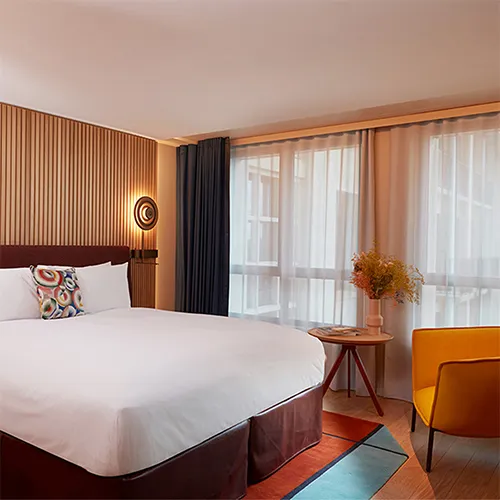 A modern hotel room with a neatly made double bed, geometric pillow, wood-paneled wall, large windows with sheer curtains, a small table with a glass vase of flowers, and a yellow armchair on a colorful rug.