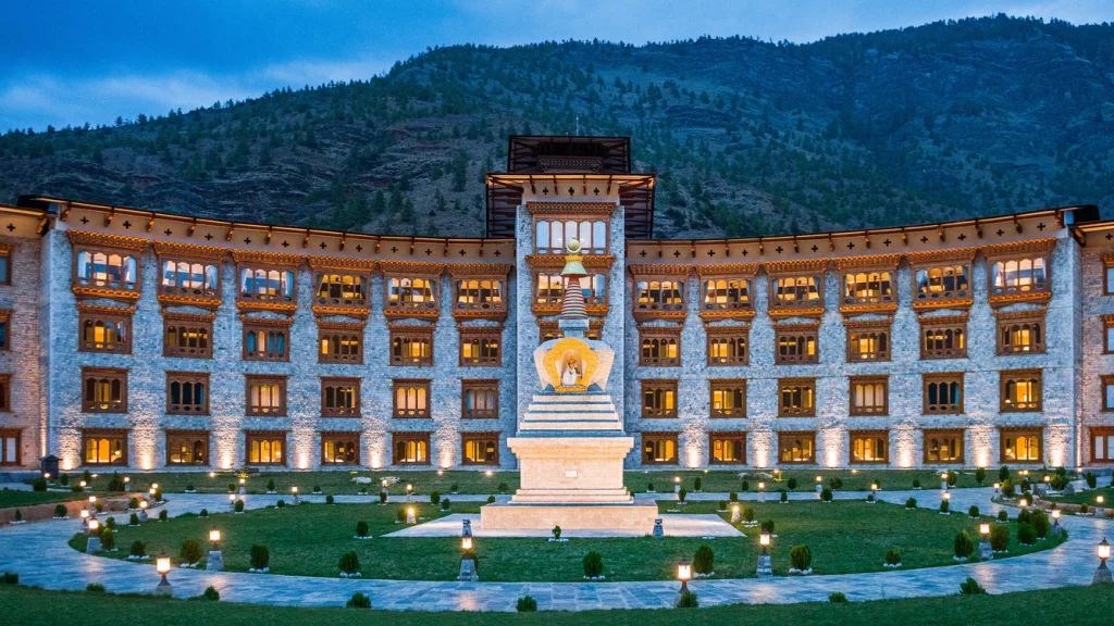 A large ornate building with traditional Bhutanese architecture, glowing warmly at dusk, stands behind a central white stupa in a landscaped courtyard with mountains in the background.