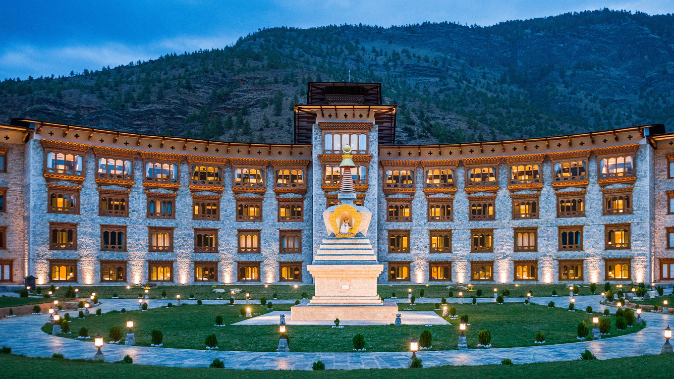 A large ornate building with traditional Bhutanese architecture, glowing warmly at dusk, stands behind a central white stupa in a landscaped courtyard with mountains in the background.
