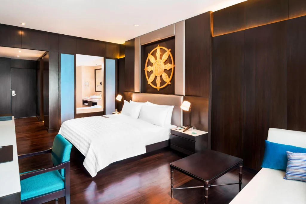 A modern hotel room with a large white bed, dark wood walls and floors, nautical wheel decor above the headboard, a teal chair, a sofa, and an open bathroom visible in the background.
