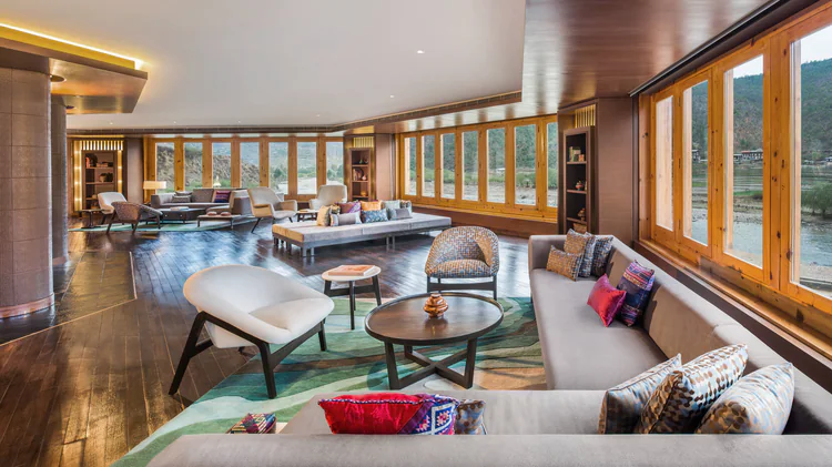 A spacious, modern lounge with large windows, wooden floors, and contemporary furniture, including sofas, armchairs, and coffee tables. Colorful pillows and natural light create a warm, inviting atmosphere.