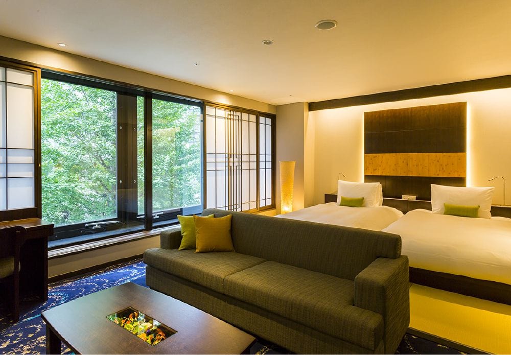 A modern hotel room with two white beds, a green-gray sofa, a desk, large windows with a view of green trees, and soft, warm lighting creating a cozy atmosphere.
