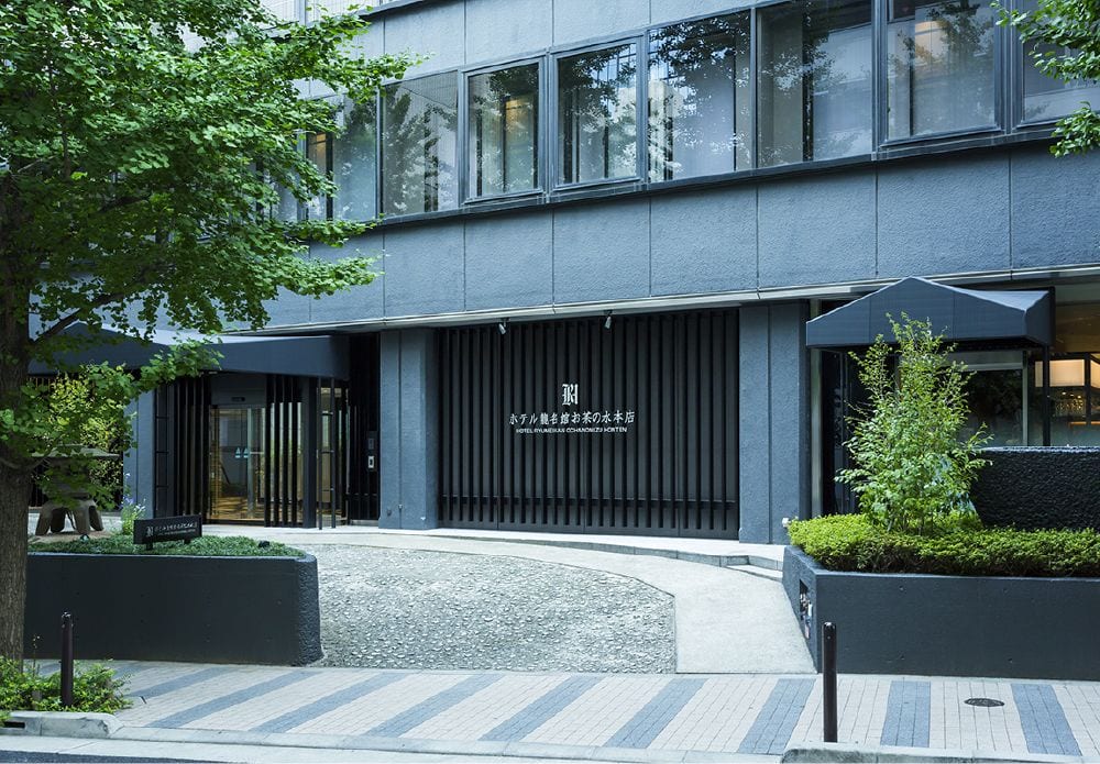 A modern gray building with large windows and a minimalist entrance, surrounded by greenery and a small landscaped courtyard with trees and bushes. Japanese text is displayed above the entrance.