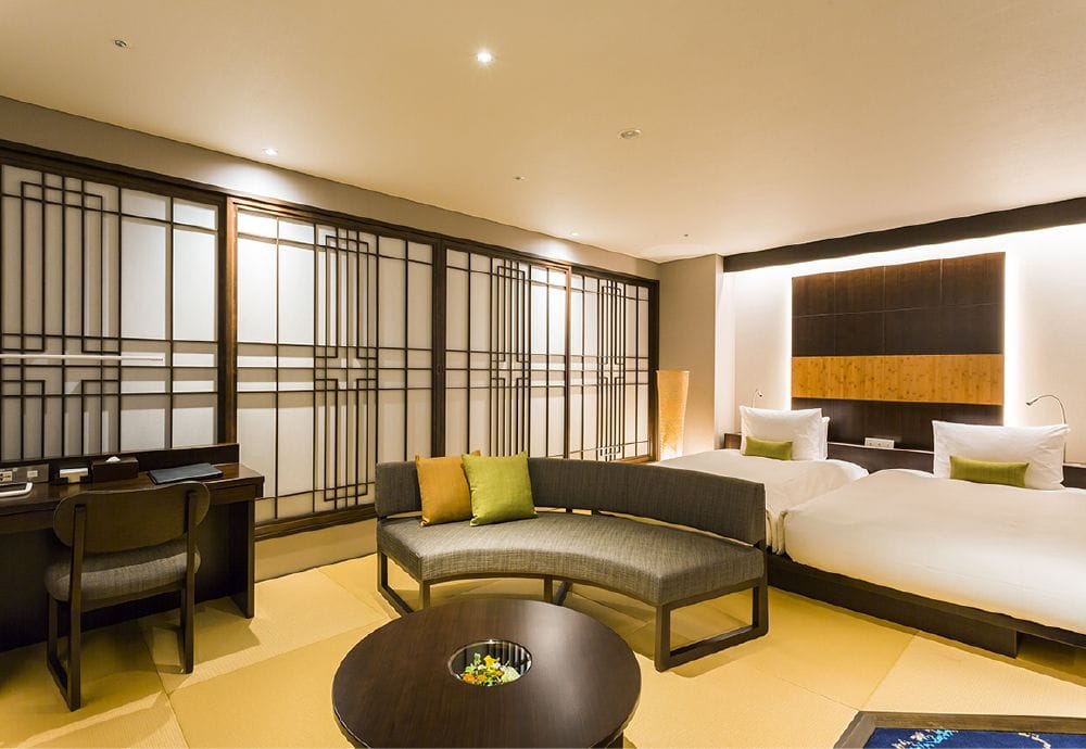 A modern Japanese-style hotel room with tatami flooring, two twin beds, a curved sofa with green and beige cushions, a round coffee table, a desk, and shoji-style sliding doors. Warm lighting creates a cozy atmosphere.