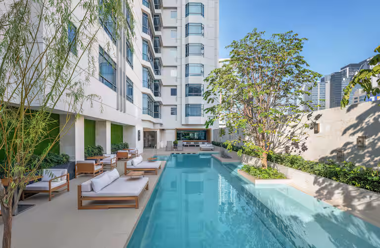 Modern outdoor swimming pool area beside a tall building, with lounge chairs, sofas, and trees providing shade; city buildings are visible in the background under a clear blue sky.