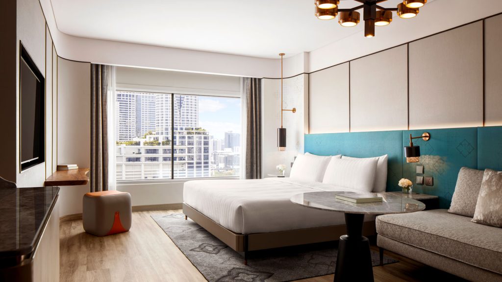 Modern hotel room with a large bed, teal headboard, small round table, gray sofa, and a window offering a cityscape view with tall buildings. Warm lighting and contemporary decor create a welcoming atmosphere.