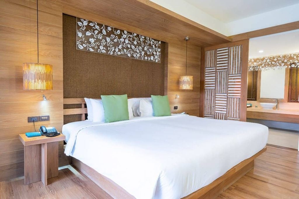 A modern hotel room with a large bed, white linens, two green pillows, wooden furniture, hanging lights, and a decorative wall panel; an open bathroom area is visible in the background.