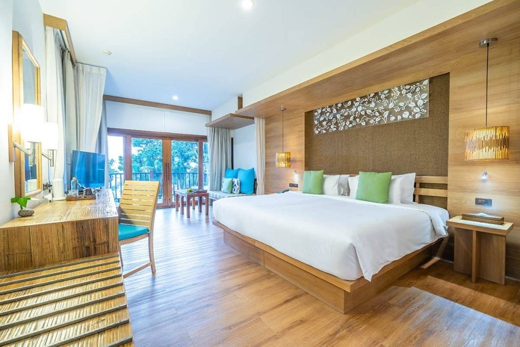 Modern hotel room with a large bed, green and blue pillows, wooden furnishings, a desk with a mirror, TV, and a seating area by floor-to-ceiling windows overlooking greenery. Natural light fills the room.
