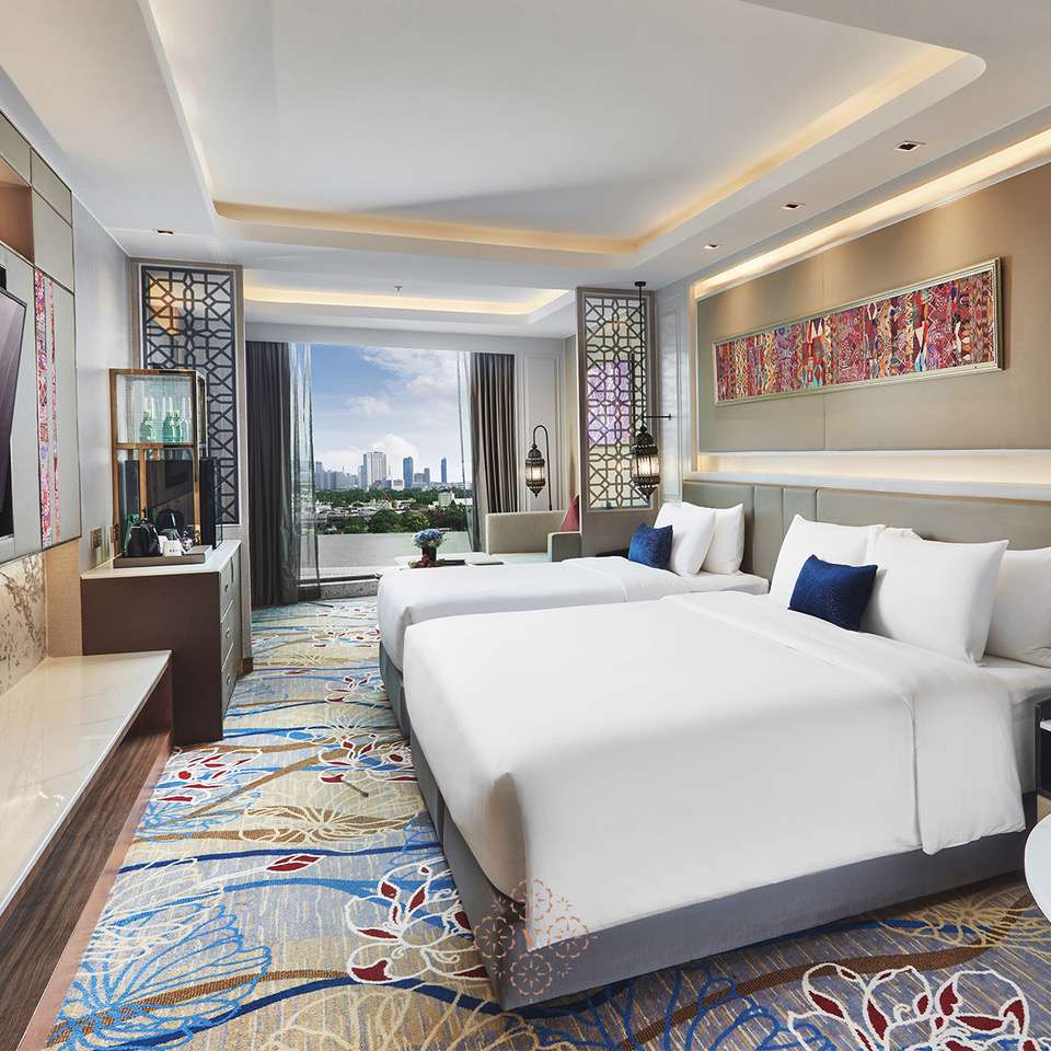 A modern hotel room with two large beds, patterned carpet, a wall-mounted TV, minibar, and a large window offering a city skyline view. The room features elegant decor with colorful artwork above the beds.