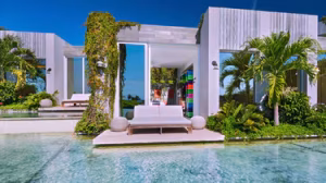 Modern villa with white exterior, open glass doors, and lush green plants. A white outdoor sofa sits on a patio above a clear, shallow pool under a bright blue sky.