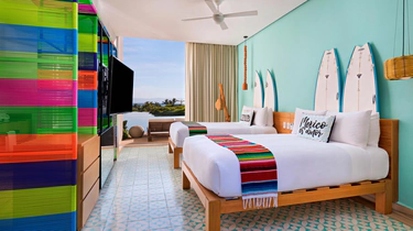 A bright hotel room with two beds, colorful striped blankets, pillows reading Nice to meet you, surfboards as wall decor, a ceiling fan, patterned tile floor, and a vibrant multicolored partition.
