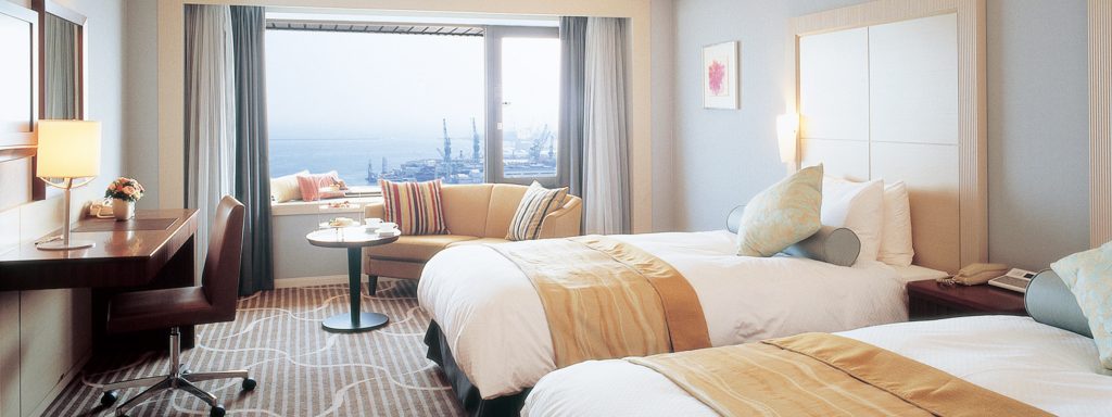 A bright hotel room with two double beds, a desk with a lamp, a small sofa by a window, and a round table with two chairs. The window offers a view of a harbor and cranes outside.