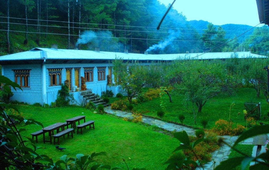 A peaceful outdoor scene shows a traditional building with ornate windows, surrounded by lush green lawns, trees, and gardens, with picnic tables, stone paths, and smoke rising in the background amidst forested hills.