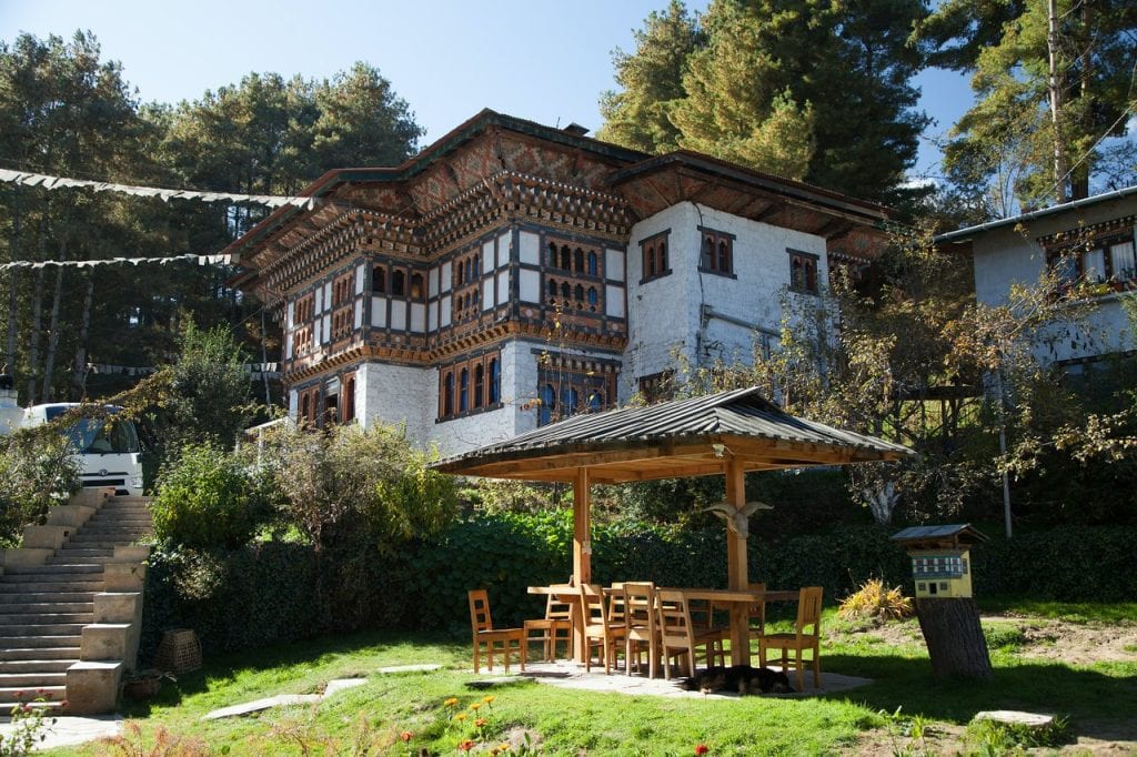 A traditional Bhutanese building with ornate wooden windows and white walls stands among trees. In the foreground, a wooden gazebo with a table and chairs sits on a grassy lawn in the sunlight.
