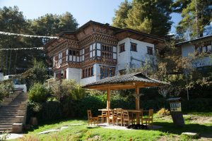 A traditional Bhutanese building with ornate wooden windows and white walls stands among trees. In the foreground, a wooden gazebo with a table and chairs sits on a grassy lawn in the sunlight.