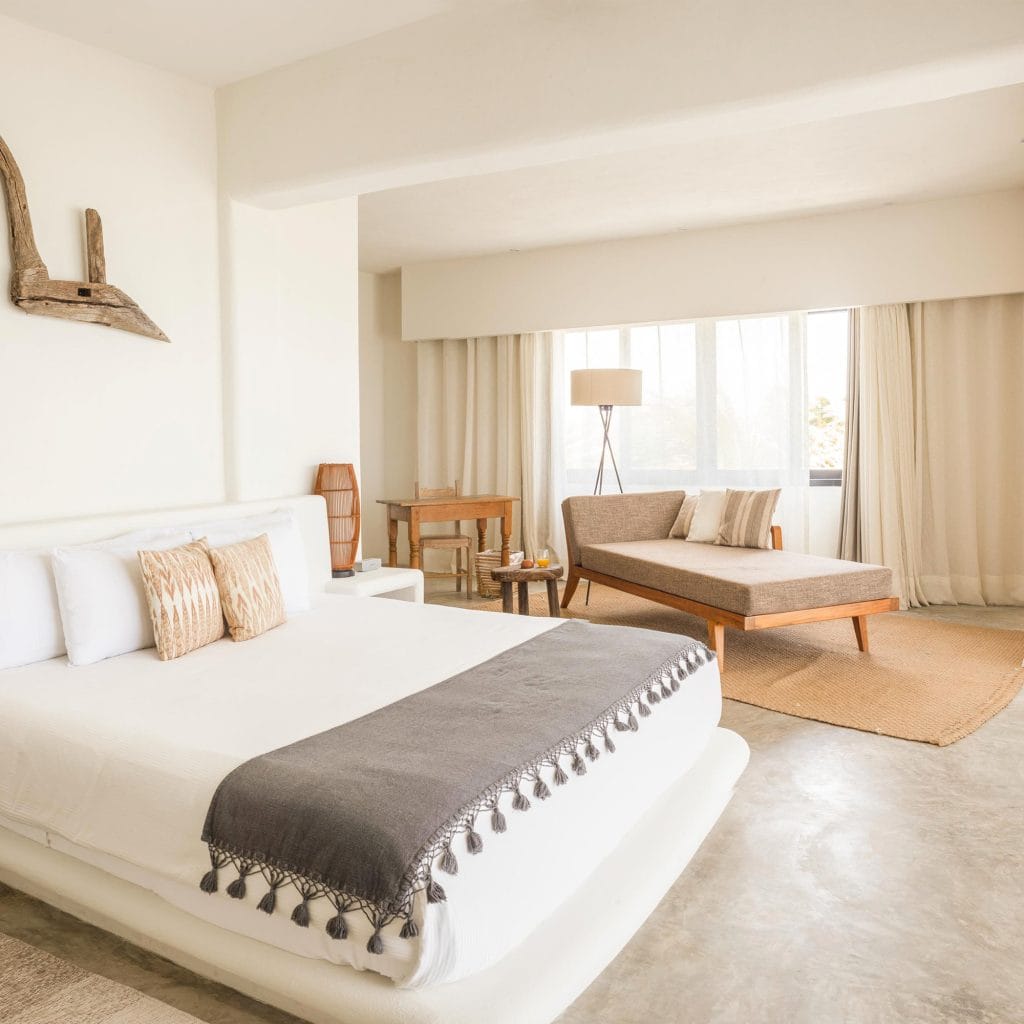 A bright, minimalist bedroom with a neatly made bed, neutral-toned pillows and blanket, a wooden lounge chair by large windows, light curtains, a rug, and a small wooden desk with a lamp.