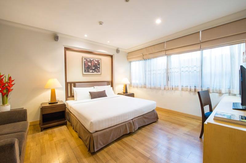 A bright, modern hotel room with a large bed, bedside tables with lamps, a sofa, desk with chair, flat-screen TV, wooden floor, and a window with beige curtains letting in natural light.