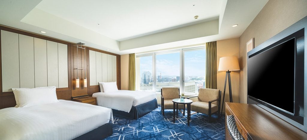 A modern hotel room with two double beds, a large window with city views, two chairs and a small table, a flat-screen TV, and warm neutral decor. Natural light brightens the space.