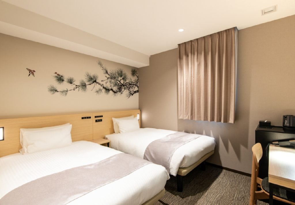 A modern hotel room with two single beds, light bedding, a desk with a chair, a kettle, and a wall decorated with a tree branch and bird mural. Beige curtains cover the window.