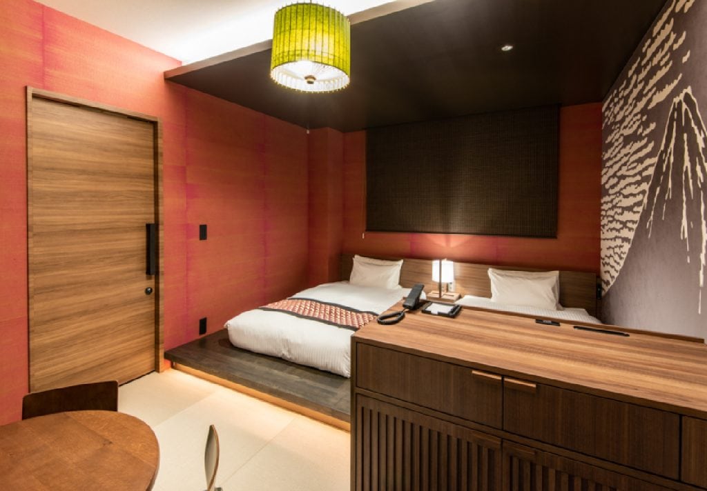 A modern hotel room with a large bed, wooden furniture, a small round table with chairs, a green hanging lamp, and red textured walls with a monochrome mural. Warm lighting creates a cozy atmosphere.