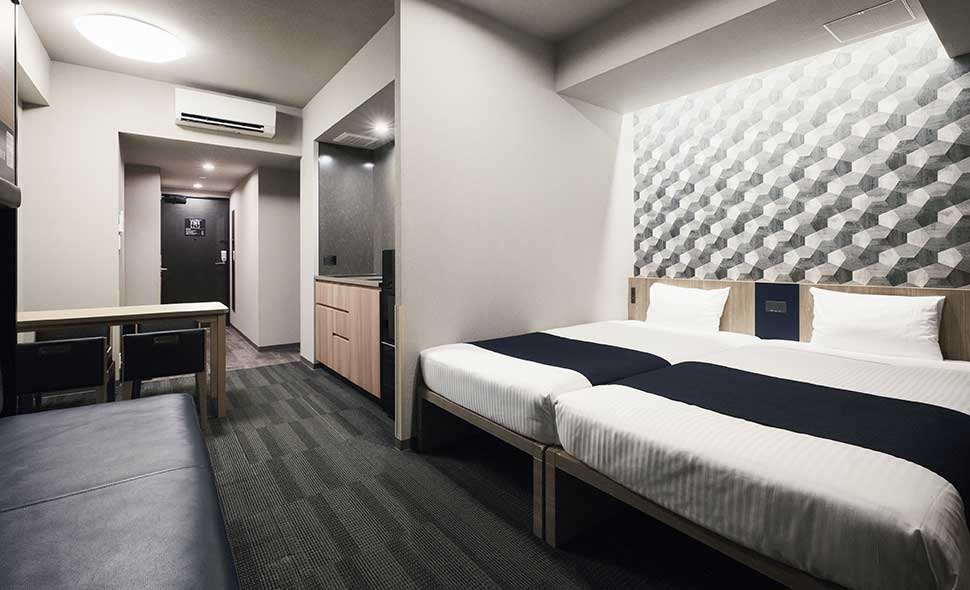 Modern hotel room with two twin beds, white bedding, and dark runners, a geometric accent wall, a small kitchenette, a seating area, and a desk, all illuminated by overhead lighting.
