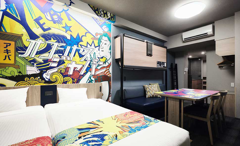 Colorful hotel room with twin beds, bright pop-art style mural on the wall, a fold-out bunk bed above a sofa, wooden table with patterned runner, and a compact kitchenette in the background.