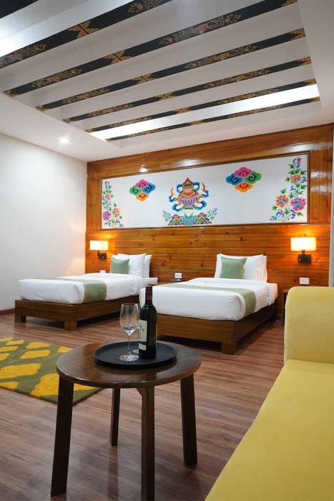 A hotel room with two single beds, colorful artwork above the headboard, modern lighting, a wooden floor, a yellow sofa, and a small table with a wine bottle and glass in the foreground.