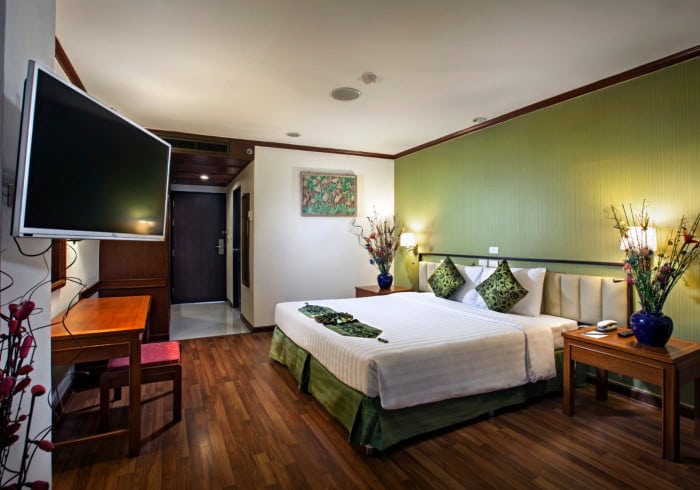 A modern hotel room with a large bed, green accent wall, two bedside tables with lamps, a flat-screen TV mounted on the wall, a wooden desk and chair, and hardwood floors. Decorative plants and artwork add color.