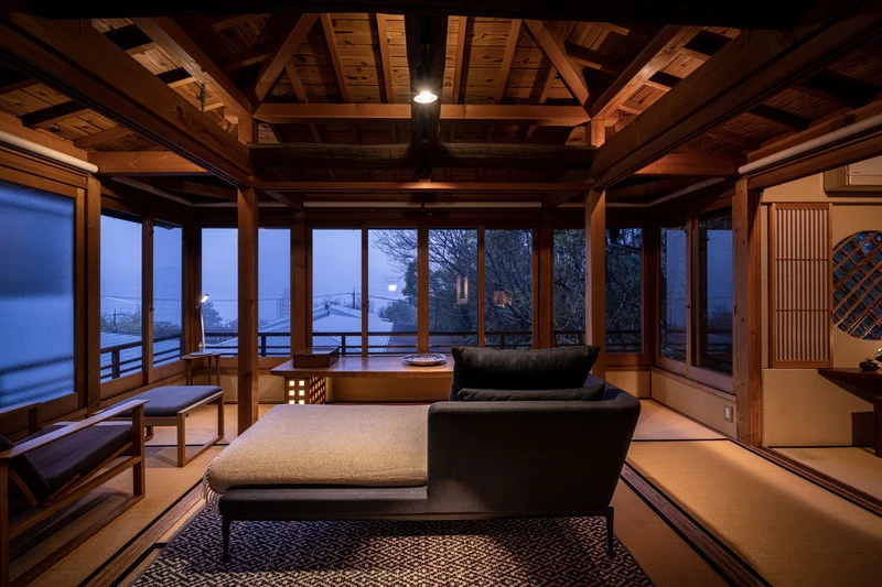 A cozy tatami room with wooden beams, large windows overlooking a misty landscape at dusk, and a modern chaise lounge in the center, blending traditional Japanese and contemporary design elements.