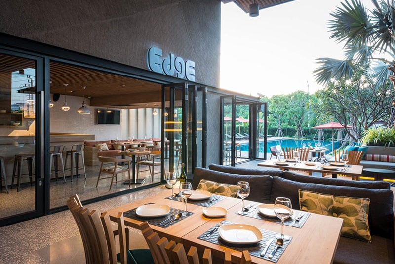 Outdoor seating area of a modern restaurant named Edge, featuring neatly set tables with plates and glasses, cushioned chairs, and a view of a pool area with trees and red umbrellas in the background.