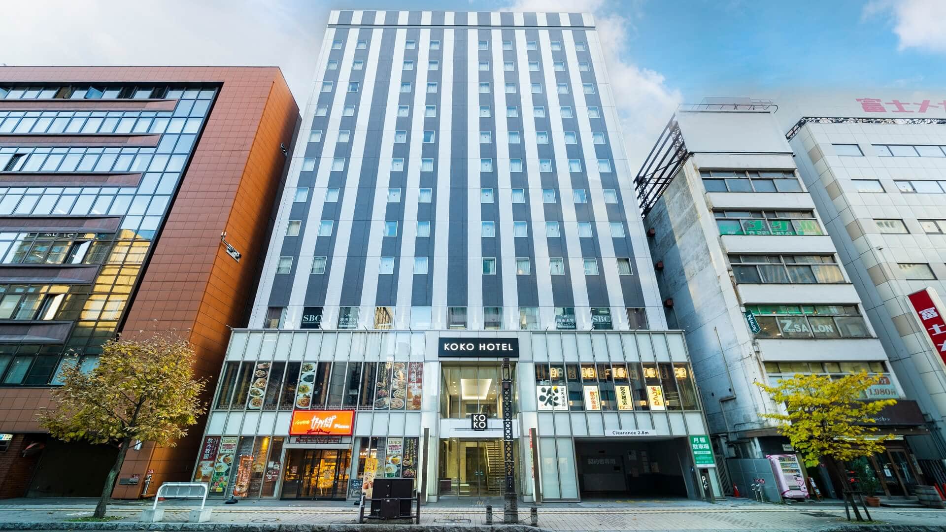 Tall modern building with striped black and white facade, labeled KOKO HOTEL at the entrance. Flanked by various other buildings, the street is lined with a few trees with yellow leaves. Bright, clear sky above.