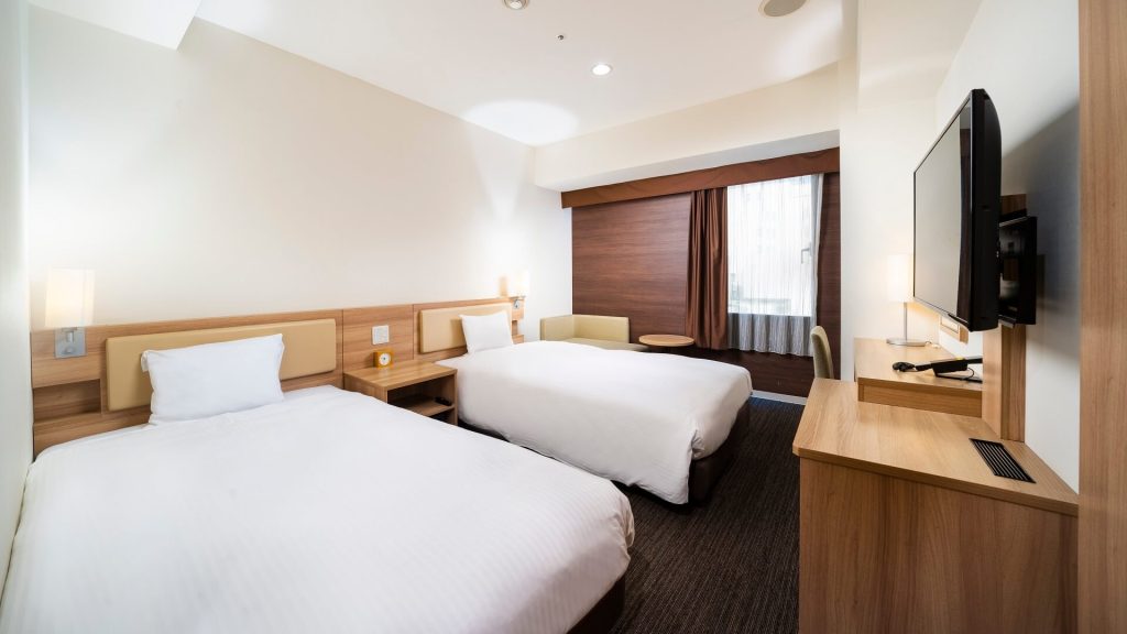A modern hotel room with two neatly made beds, a wall-mounted TV, a wooden desk, a chair by the window, and soft lighting creating a bright, clean, and comfortable atmosphere.