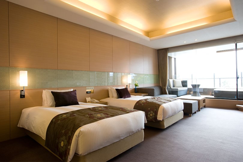 A modern hotel room with two twin beds, soft lighting, wood-paneled walls, a sitting area with a sofa, a large window, and a balcony with outdoor seating.
