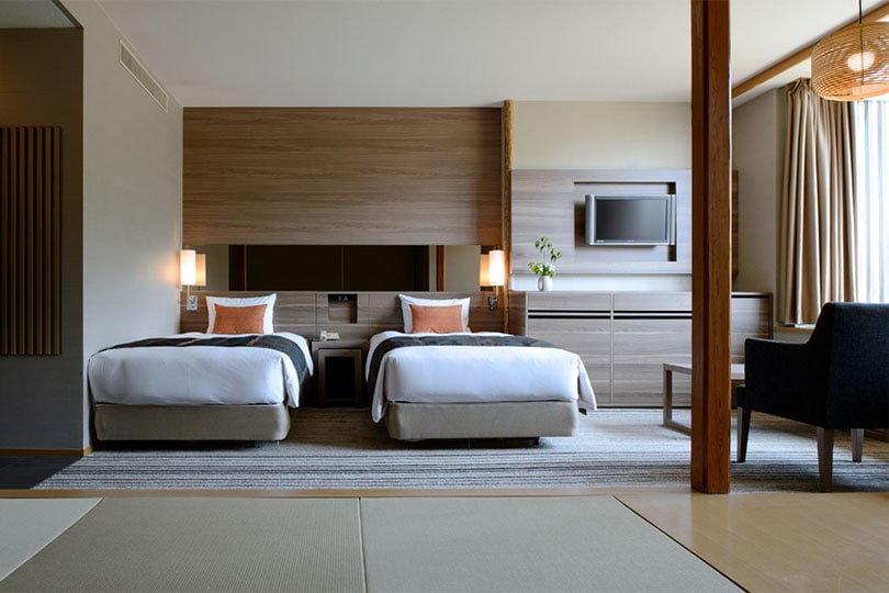 Modern hotel room with two twin beds, orange accent pillows, a wall-mounted TV, nightstand, chair, table, large window with curtains, and minimalistic wooden decor. Natural light fills the room.