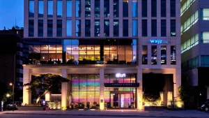 Exterior view of a modern, multi-story hotel building at dusk, with large glass windows lit up in warm colors. The hotel entrance is illuminated, and aloft and w xyz lounge signs are visible.