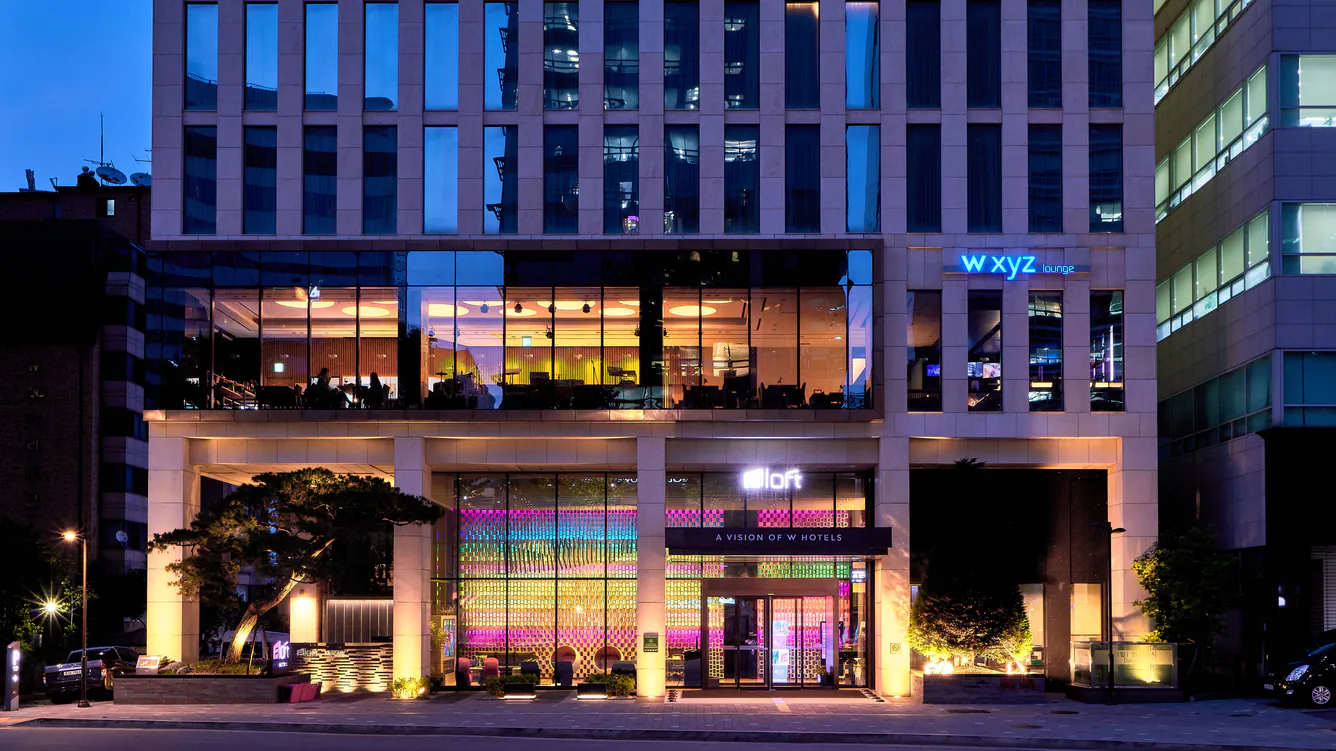 Exterior view of a modern, multi-story hotel building at dusk, with large glass windows lit up in warm colors. The hotel entrance is illuminated, and aloft and w xyz lounge signs are visible.