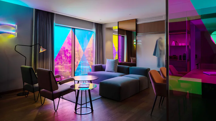 A modern hotel suite with a sofa, chairs, coffee table, and large windows. Colorful neon lighting in pink, yellow, and blue creates a vibrant, artistic ambiance throughout the room.