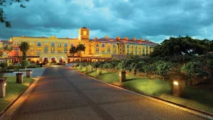 A large, elegant yellow hotel with warm lights illuminating its facade at dusk, surrounded by manicured lawns, trees, and a paved driveway leading to the entrance under a dramatic cloudy sky.