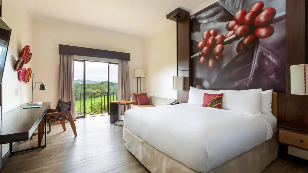 A modern hotel room with a large bed, a mural of coffee cherries above the headboard, desk and chair, armchair by the window, and a balcony with a scenic view of greenery and hills outside.