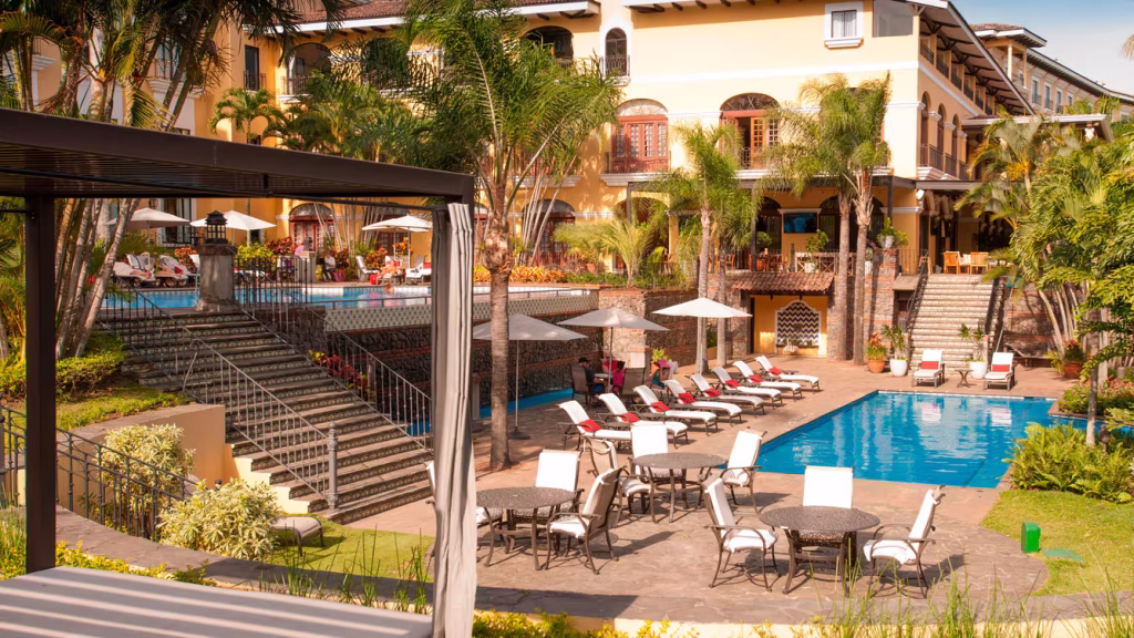 A luxurious hotel with yellow walls overlooks a garden area with a rectangular swimming pool, sun loungers, umbrellas, and tropical plants. Stairs connect the pool area to a terrace with more seating and palm trees.