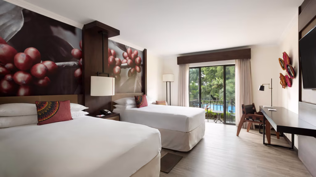 Modern hotel room with two large beds, a desk, a flat-screen TV, and a large window overlooking a pool and greenery. The room features contemporary decor and a vibrant mural of red berries above the beds.