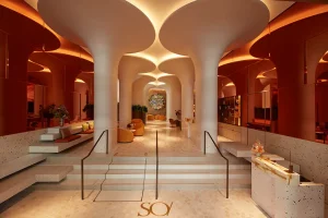A modern, stylish hotel lobby with sculptural, tree-like columns, warm ambient lighting, curved seating areas, and terrazzo floors. The space feels luxurious and inviting, with a glowing ceiling and elegant decor.