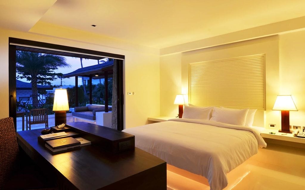 Modern hotel room with a large bed, two bedside lamps, a work desk, and an open sliding door leading to a patio with outdoor seating and a view of trees in the evening light.