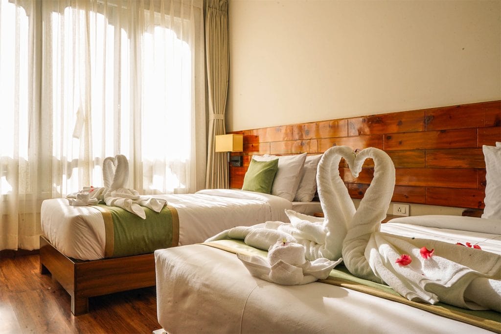 A bright hotel room with two beds, each decorated with towels folded into swan shapes forming hearts. Pink flower petals are scattered on the beds, and sunlight streams through sheer curtains.