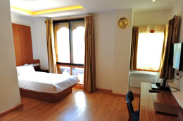 A modern hotel room with wooden floors, a double bed near a large window with gold curtains, a desk and chair, a wall-mounted TV, and a clock on the wall. The room is bright with natural light.