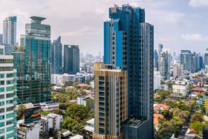 A cityscape featuring tall modern skyscrapers, including a prominent blue and gold high-rise in the foreground, surrounded by other glass buildings and greenery under a partly cloudy sky.