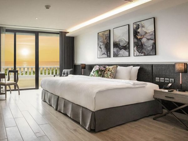 Modern hotel room with a large bed, abstract artwork above the headboard, a small table with chairs near glass doors, and a balcony overlooking a sunset view of the ocean. Warm lighting and neutral decor create a cozy atmosphere.