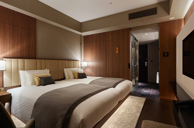 A modern hotel room with two large beds, neutral bedding, wooden paneling, bedside lamps, a soft rug, and a glass-walled bathroom in the background. The lighting is warm and inviting.
