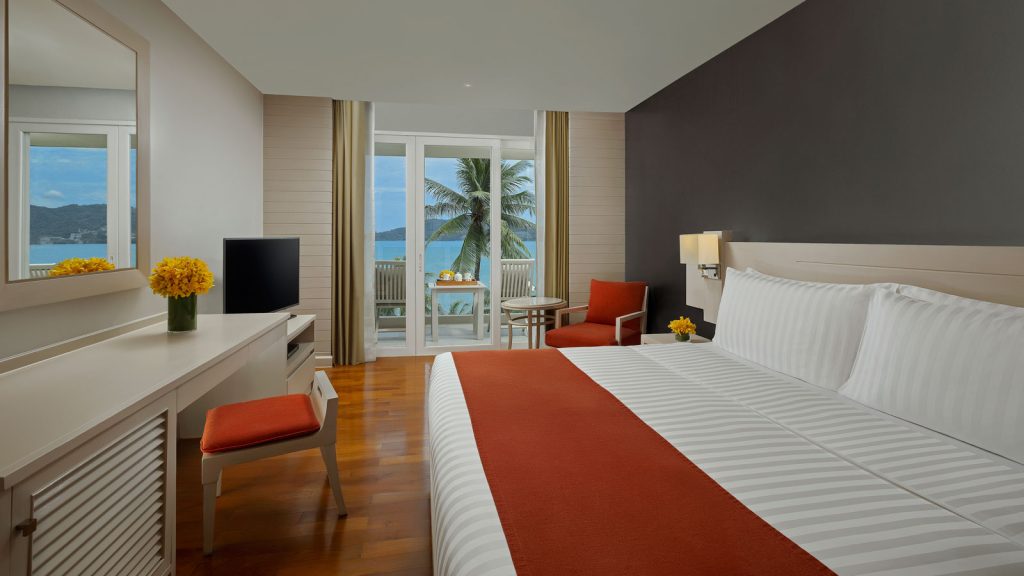 A modern hotel room with a large bed featuring white and orange bedding, a desk with a chair, a TV, and a balcony overlooking the ocean, palm trees, and mountains. Yellow flowers decorate the desk and side table.